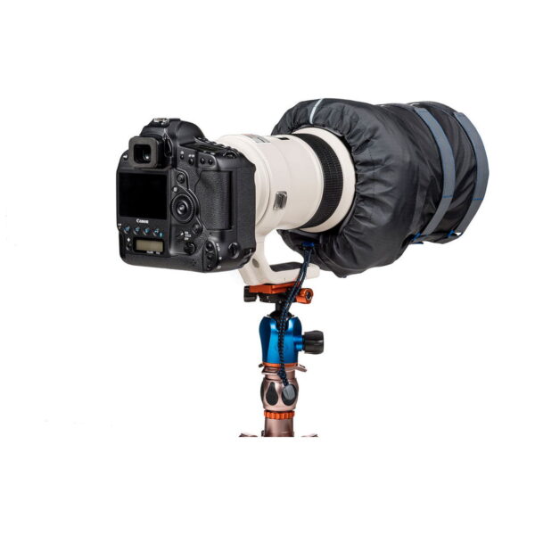 Think Tank Hydrophobia DM 300-600 V3 musta sadesuoja peittää ammattikäyttöön tarkoitetun DSLR-kameran, jossa on suuri teleobjektiivi ja joka on asennettu jalustalle, jossa on sinisiä ja kuparisia korostuksia valkoista taustaa vasten.