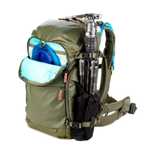 The Shimoda Designs Explore v2 25 Starter Kit Camera Backpack, green, features a partially opened front pocket with a blue hydration bladder, a blue tube on the shoulder strap and a camera stand attached to the side.
