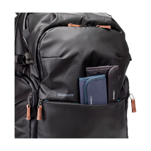 Close-up of a black Shimoda Designs Explore v2 30 camera backpack with a partially open front zippered pocket and two padded cases (one blue and one grey). Features include brown zipper pulls and a subtle grid pattern on the fabric.