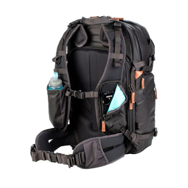The Shimoda Designs Explore v2 30 camera backpack, black, features a white base with padded shoulder straps, multiple pockets and a hipbelt; a water bottle and smartphone are stored in the side and hipbelt pockets.