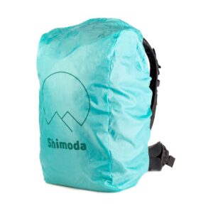 Turkoosi Shimoda-logolla varustettu sadesuojus sopii hyvin vihreän Shimoda Designs Explore v2 30 -kamerareppu -mallin päälle, ja oikealla puolella näkyy osia olkahihnasta ja lantiovyöstä.