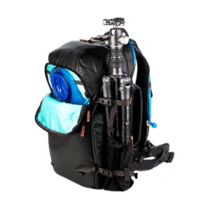 The Shimoda Designs Explore v2 35 Camera Backpack, Black features an open side pocket revealing a blue water tank with a tripod attached to the outside and blue hydration hoses extending from the backpack.