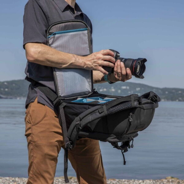Henkilö seisoo veden äärellä pitelemässä kameraa, yllään Tenba Axis V2 16L Backpack -kamerareppu, jonka avoin etuläppä paljastaa sen järjestetyn sisätilan kameravarusteille. Reppu on kiinnitetty, jotta siihen pääsee helposti käsiksi.