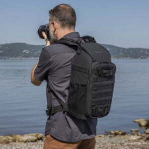 Parrakas mies tummassa paidassa ja ruskeissa housuissa seisoo järven rannalla, taustalla kukkulat, ottaa valokuvia ja kantaa Tenba Axis V2 16L Backpack -kamerareppua.