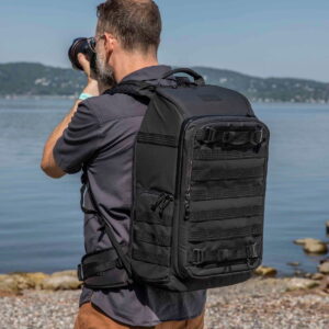 Lyhythiuksinen ja parrakas mies, jolla on tumma paita ja mukanaan Tenba Axis V2 24L Backpack -kamerareppu, seisoo kivikkoisella rannalla ja ottaa valokuvia. Taustalla näkyy vettä ja kaukaisia kukkuloita.
