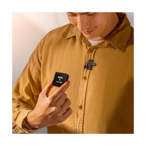 A man in a tan shirt clips the Rode FlexClip GO microphone to his collar, holds the sleek black wireless transmitter in his hand and smiles as he looks down at the device.