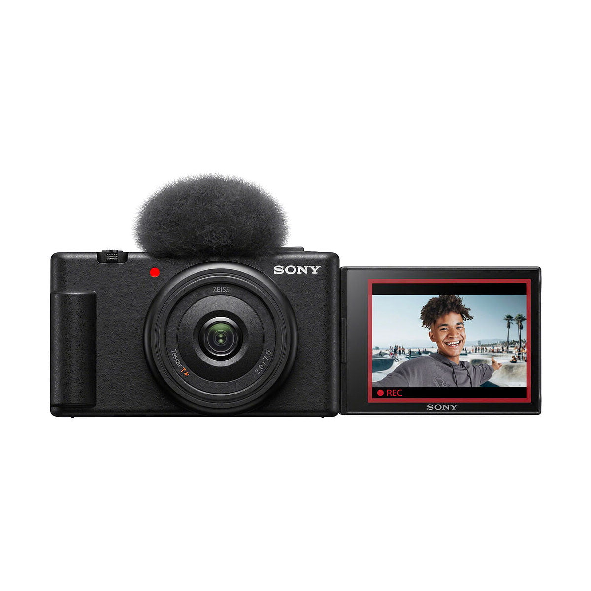 The black Sony ZV-1F compact camera with flip-out screen shows a smiling person being filmed outdoors, with a fuzzy microphone attached.