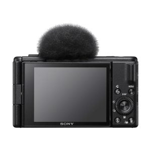 Kuvassa näkyy takana musta Sony ZV-1F -kompaktikamera, jossa on suuri LCD-näyttö ja ohjauspainikkeet, sekä sumea mikrofoni tuulisuoja kiinnitetty yläosaan.