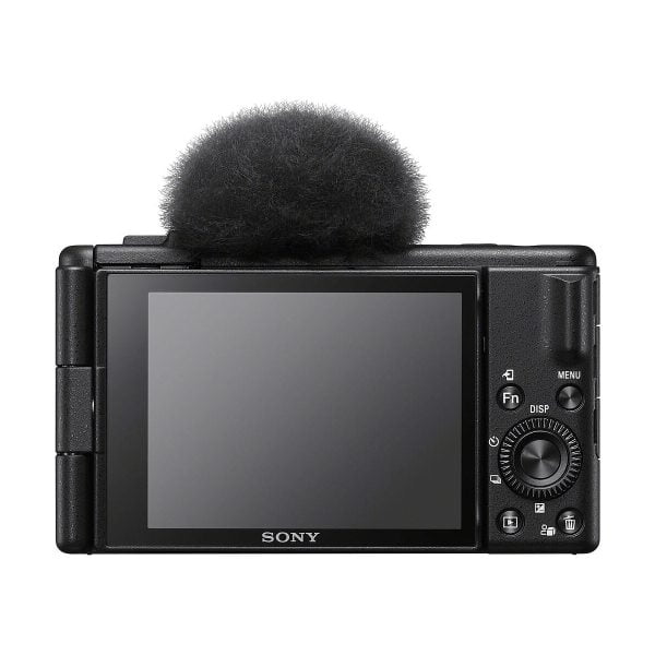Kuvassa näkyy takana musta Sony ZV-1F -kompaktikamera, jossa on suuri LCD-näyttö ja ohjauspainikkeet, sekä sumea mikrofoni tuulisuoja kiinnitetty yläosaan.