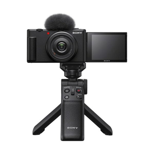 Musta Sony ZV-1F -kompaktikamera, jossa on ulosvedettävä näyttö ja yläosan mikrofoni, asennettuna kompaktin Sonyn jalustakahvan varaan, valkoista taustaa vasten.