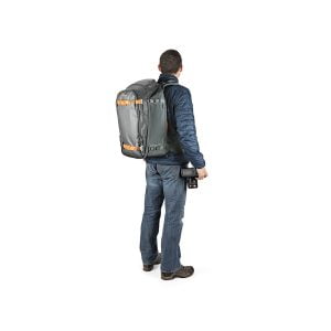 Siniseen takkiin ja farkkuihin pukeutunut mies seisoo kasvot menosuuntaan, hänellä on Lowepro Whistler BP 350 AW II GL -kamerarepussa oranssit korostukset ja hänellä on kamera oikeassa kädessään.