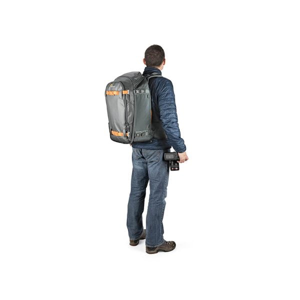 Siniseen takkiin ja farkkuihin pukeutunut mies seisoo kasvot menosuuntaan, hänellä on Lowepro Whistler BP 350 AW II GL -kamerarepussa oranssit korostukset ja hänellä on kamera oikeassa kädessään.