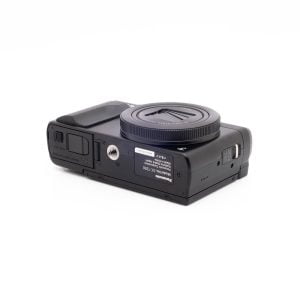 Panasonic DC-TZ90 7657-1 Panasonic Lumix DC-TZ90 - Käytetty kompakti musta digitaalikamera makaa selällään, jolloin sen alapuoli, jossa on tarrat, jalustakiinnike, akkulokero ja muistikorttipaikka, paljastuu valkoista taustaa vasten.