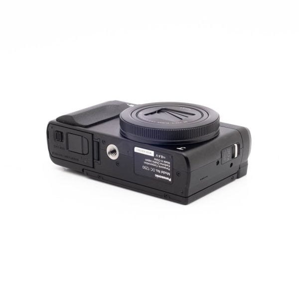 Panasonic DC-TZ90 7657-1 Panasonic Lumix DC-TZ90 - Käytetty kompakti musta digitaalikamera makaa selällään, jolloin sen alapuoli, jossa on tarrat, jalustakiinnike, akkulokero ja muistikorttipaikka, paljastuu valkoista taustaa vasten.