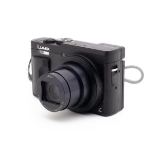 Panasonic DC-TZ90 7657-4 Panasonic Lumix DC-TZ90 - Käytetty digitaalikamera on kuvattu vasemmalle kallistettuna valkoisella taustalla, ja siihen on kiinnitetty osittain laajennettu Leica-zoom-objektiivi ja harmaa rannehihna.