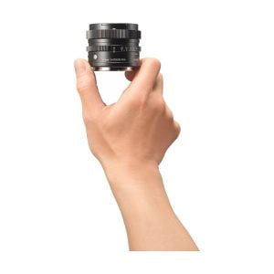Käsi pitää Sigma 17mm f/4 DG DN DN Contemporary -objektiivia Sonylle pystyssä valkoista taustaa vasten. Mustassa objektiivissa on näkyvät merkinnät ja säätörenkaat, ja käsi näkyy sivulta.