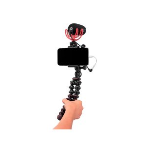 Käsi pitää JOBY GripTight PRO 2 GorillaPod -jalustaa älypuhelimille, johon on kiinnitetty puhelin, ulkoinen mikrofoni ja kaapeli, valmiina mobiilitallennukseen tai vloggaukseen valkoisella taustalla.