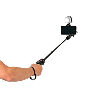 Joby-Beamo-Mini-6 Henkilö pitää kädessään selfiekeppiä, johon on kiinnitetty älypuhelin, kiinnitetty Joby Beamo Mini LED-valo ja rannehihna. Keppi on ojennettu ja puhelin on henkilöstä poispäin.