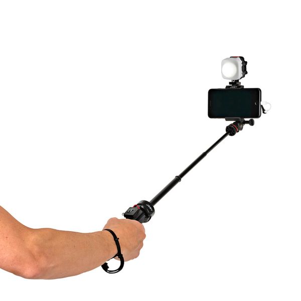 Joby-Beamo-Mini-6 Henkilö pitää kädessään selfiekeppiä, johon on kiinnitetty älypuhelin, kiinnitetty Joby Beamo Mini LED-valo ja rannehihna. Keppi on ojennettu ja puhelin on henkilöstä poispäin.