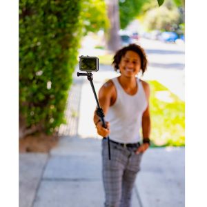 Henkilö seisoo aurinkoisella jalkakäytävällä ja hymyilee kameraan, jota pidetään Joby Compact 2in1 Monopod -selfiekepillä. Puut ja viherkasvit korostavat kirkasta ulkoilmataustaa.