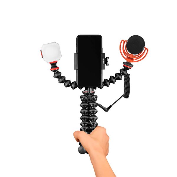 Käsi pitää Joby GorillaPod Advanced Mobile Vlogging Kit - jossa on asennettu älypuhelin, LED-valo vasemmalla ja ulkoinen mikrofoni punaisella iskukiinnikkeellä oikealla - valkoisella taustalla.