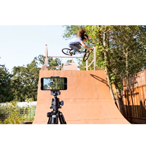 Henkilö tekee BMX-temppua puisella rampilla ulkona, kun Joby GripTight Pro Phone Mount -puhelinpidikkeeseen kiinnitetty älypuhelin, joka on jalustalla, tallentaa toimintaa puiden ja puuaidan taustalla.