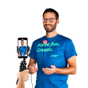 A smiling man with glasses and a blue "Have fun. Create." t-shirt standing in front of a white background, photographed with a smartphone on a flexible tripod using the Joby Wavo Lav Microphone.