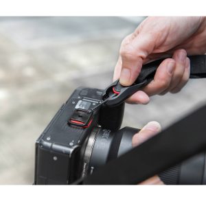 Lähikuva henkilöstä, joka kiinnittää Peak Design Anchor Mount -kiinnikkeen DSLR-kameraan pikakiinnikkeellä. Tarkennus on käsissä ja kamerassa, ja ulkotilojen tausta on pehmeästi sumea.