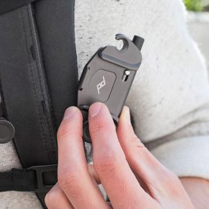 The person attaches the Peak Design Capture (Clip Only) - Black, a small grey metallic clip with a logo to the strap of a black backpack while wearing a light grey shirt.