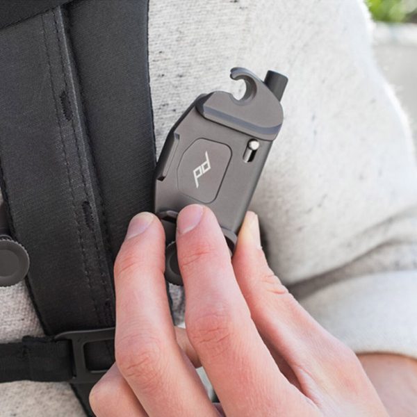 The person attaches the Peak Design Capture (Clip Only) - Black, a small grey metallic clip with a logo to the strap of a black backpack while wearing a light grey shirt.