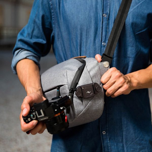 A person wearing a blue button-down shirt is holding a camera in one hand and a grey shoulder bag with a Peak Design Capture (Clip Only) - Black attached to the bag strap across the chest.