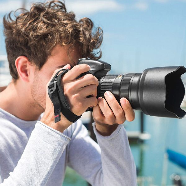 A person with curly hair and a light grey shirt wears a DSLR camera with a Peak Design Clutch strap as they prepare to take photos out on the water on a sunny day.