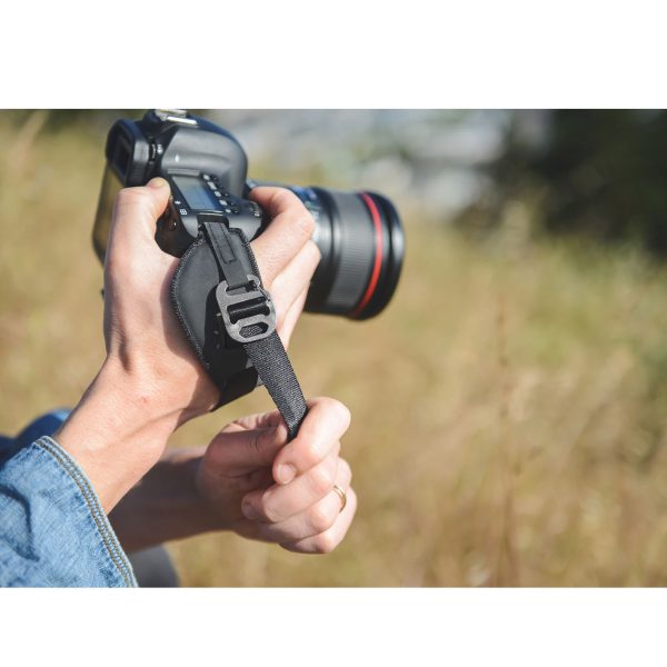 A person in jeans holds a DSLR camera with both hands and adjusts the strap of the Peak Design Clutch strap outside on the sunny lawn.
