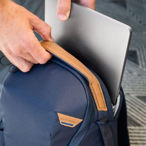 Henkilö laittaa hopeanvärisen kannettavan tietokoneen tummansinisen Peak Design Everyday Backpack Zip 20L -kamerareppu -repun pehmustettuun lokeroon, jossa on ruskehtava nahkakahva ja joka lepää kuviollisella matolla.