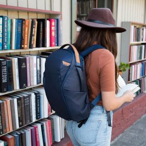 Ruskeaan hattuun, ruskeaan paitaan ja farkkuihin pukeutunut nainen, jolla on Peak Design Everyday Backpack Zip 20L -kamerareppu, tummansininen selailee ulkona olevaa kirjahyllyä, kädessään avoin kirja.