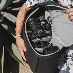 Kukkaiseen paitaan pukeutunut henkilö, jolla on tatuoitu käsivarsi, pitää mustaa Peak Design Everyday Backpack v2 30L -reppua auki ja näyttää DSLR-kameran ja useita objektiiveja siististi järjestettynä sen pehmustetuissa lokeroissa.