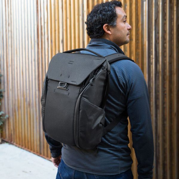 Tummiin takkeihin pukeutunut mies, jolla on mukanaan Peak Design Everyday Backpack v2 30L -kamerareppu, musta, seisoo ulkona pystysuoran puuaidan vieressä ja katsoo oikealle.