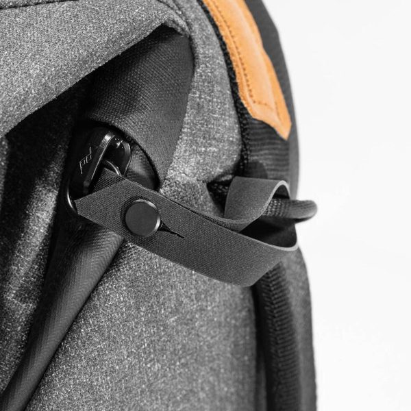 Lähikuva Peak Design Everyday Backpack v2 30L -repusta tummanharmaana, jossa on musta hihna nappikiinnityksellä ja pieni ruskehtava nahkainen korostus oikeassa yläkulmassa. Kuvioidut materiaalit näyttävät kestäviltä.