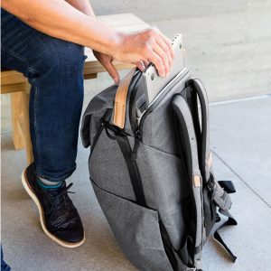 Farkkuihin ja mustiin lenkkareihin pukeutunut henkilö istuu penkillä ja laittaa kannettavan tietokoneen tummanharmaa Peak Design Everyday Backpack v2 30L -kamerareppu lepää maassa vieressä.