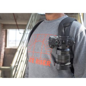 Harmaaseen graafiseen collegepaitaan pukeutunut henkilö on sisätiloissa portaiden ja ikkunan lähellä, ja hänellä on mukanaan Sony-kamera, joka on kiinnitetty Peak Design Everyday Backpack v2 30L (tummanharmaa) -repun hihnaan.