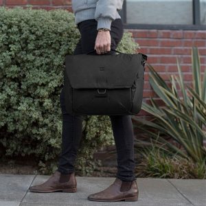 Peak-Design-Everyday-Messenger-13L-v2---Black-7 Tummiin housuihin, ruskeisiin saappaisiin ja vaaleanharmaaseen villapaitaan pukeutunut henkilö kävelee jalkakäytävällä pitäen kädessään Peak Design Everyday Messenger 13L V2 - Black. Taustalla näkyy vihreitä pensaita ja tiiliseinä.
