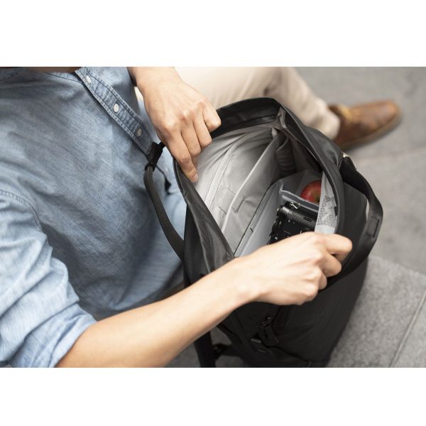 Vaaleansiniseen paitaan pukeutunut henkilö avaa mustan Peak Design Everyday Totepack 20L v2 -repun, jonka sisältä paljastuu puhelin, kamera ja omena harmaalla alustalla istuen. Vain ylävartalo ja kädet ovat näkyvissä.