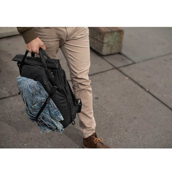 Khakihousuihin ja ruskeisiin kenkiin pukeutunut henkilö kävelee jalkakäytävällä, mukanaan Peak Design Everyday Totepack 20L v2 - Black, johon on kiinnitetty farkkutakki. Vain hänen alavartalonsa ja kätensä ovat näkyvissä.