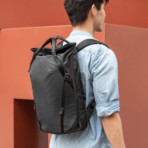 Henkilö, jolla on vaaleansininen farkkupaita ja mukanaan Peak Design Everyday Totepack 20L v2 - Black, seisoo modernin, ruosteenvärisen seinän edessä kamerasta poispäin.