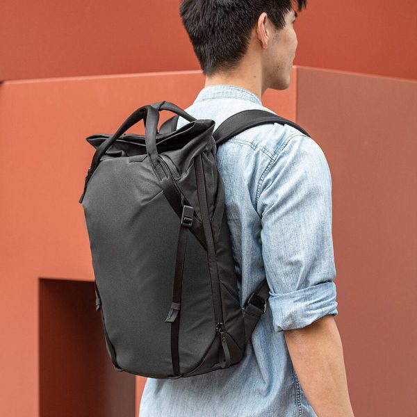 Henkilö, jolla on vaaleansininen farkkupaita ja mukanaan Peak Design Everyday Totepack 20L v2 - Black, seisoo modernin, ruosteenvärisen seinän edessä kamerasta poispäin.