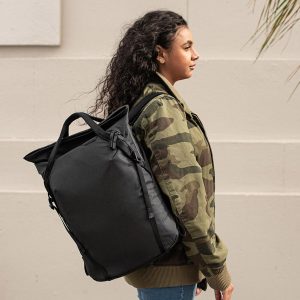 Kiharatukkainen nainen naamiotakissa seisoo sivuttain vaaleaa seinää vasten, selässään musta Peak Design Everyday Totepack 20L v2.