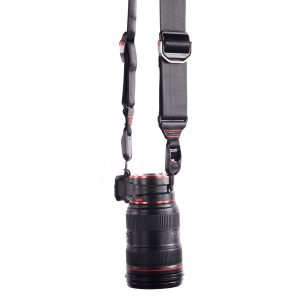 Peak-Design-Lens-Kit-Canon-4 Peak Design Lens Kit - Canon EF, jossa on musta objektiivi punaisilla korostuksilla, on kiinnitetty pystysuoraan leveään, säädettävään mustaan hihnaan, jossa on metalliklipsit ja -kiinnikkeet, valkoista taustaa vasten.