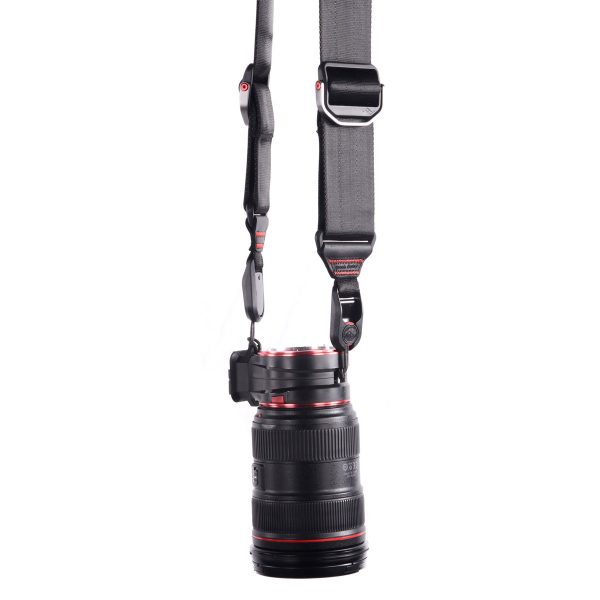 Peak-Design-Lens-Kit-Canon-4 Peak Design Lens Kit - Canon EF, jossa on musta objektiivi punaisilla korostuksilla, on kiinnitetty pystysuoraan leveään, säädettävään mustaan hihnaan, jossa on metalliklipsit ja -kiinnikkeet, valkoista taustaa vasten.