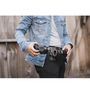 Peak-Design-Lens-Kit-Canon-5 Farkkutakki päällä ja puisen aidan edessä seisova henkilö vaihtaa Canon EF -objektiiveja Peak Design Lens Kit -objektiivisarjaan, jokainen objektiivi kädessä, samalla kun hänellä on vyötärönsä ympärillä kamerakotelovyö.