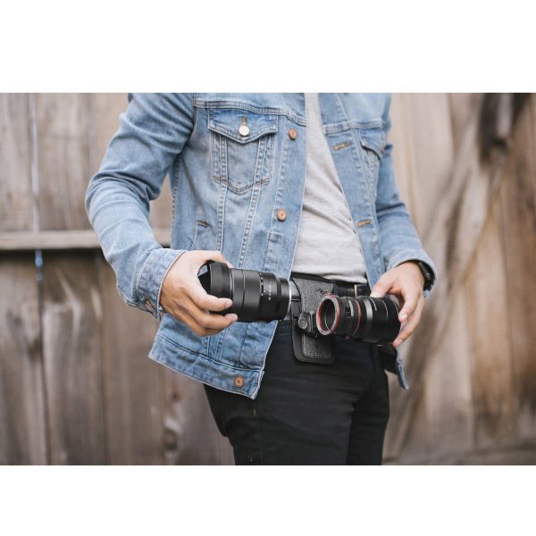 Peak-Design-Lens-Kit-Canon-5 Farkkutakki päällä ja puisen aidan edessä seisova henkilö vaihtaa Canon EF -objektiiveja Peak Design Lens Kit -objektiivisarjaan, jokainen objektiivi kädessä, samalla kun hänellä on vyötärönsä ympärillä kamerakotelovyö.